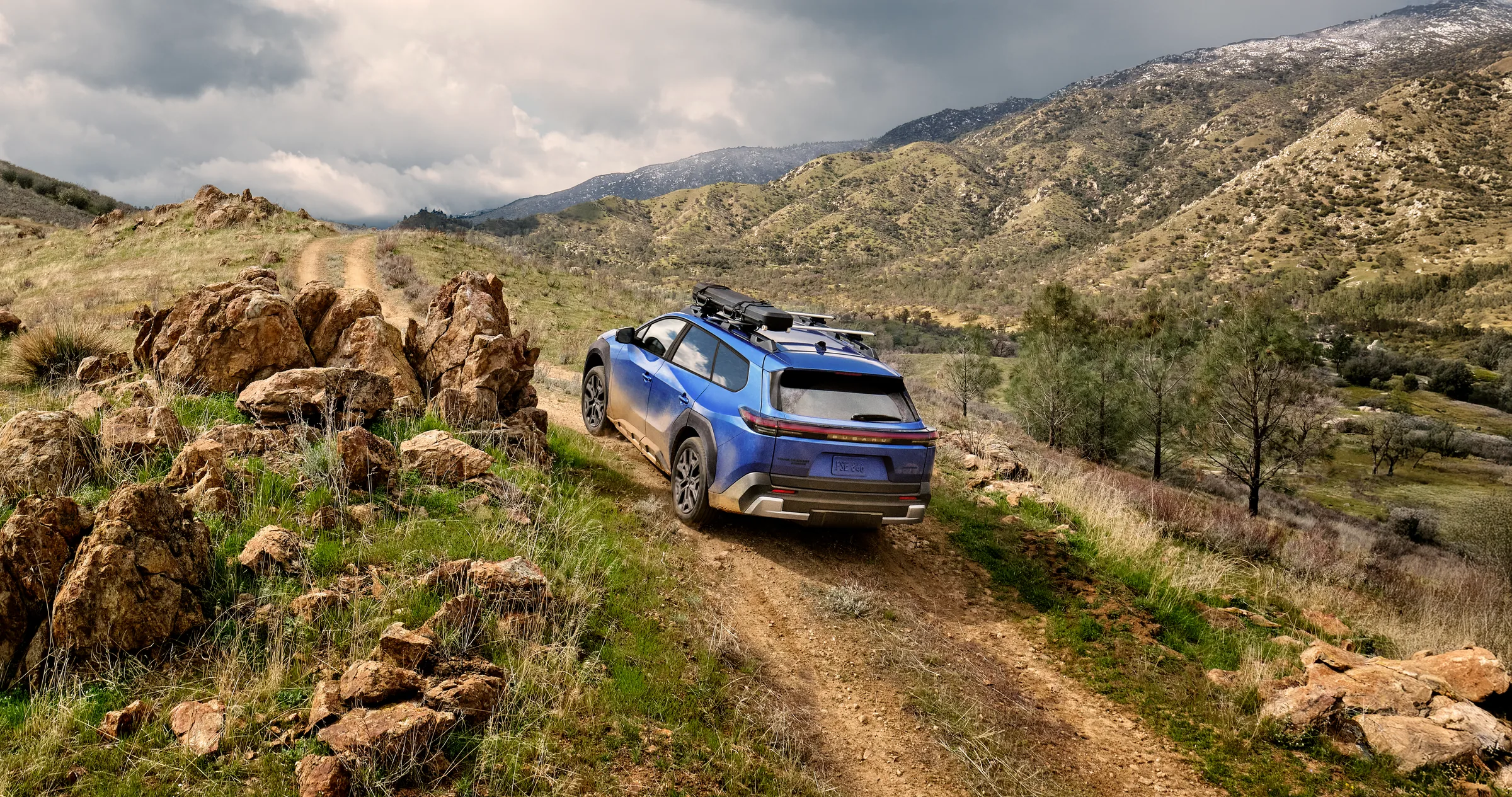 A static image of a blue 2026 Subaru Trailseeker driving on a narrow dirt trail that winds through rocky, grassy hills. 