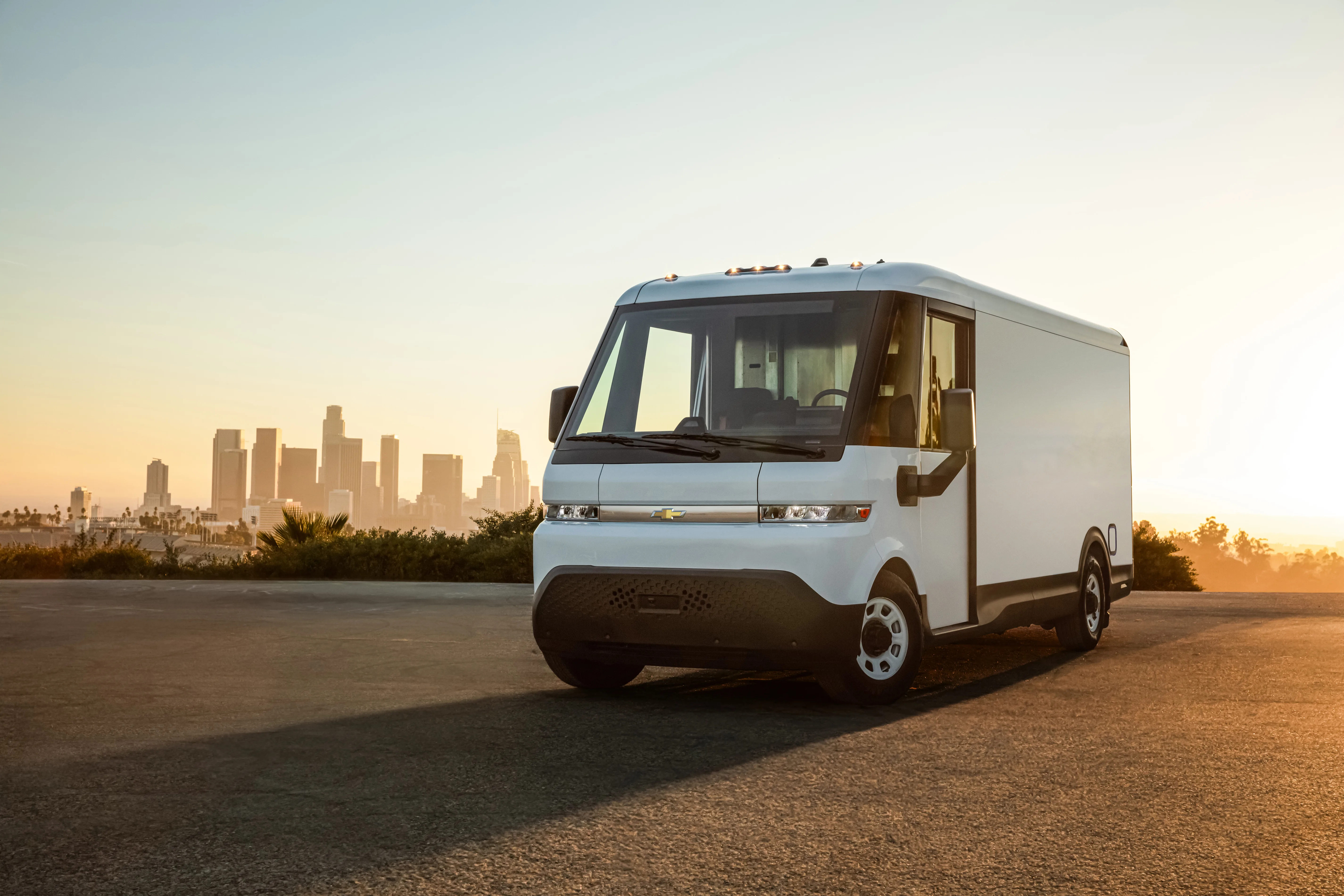 A white Chevrolet electric step van parked on a paved overlook at sunset, with a legible Chevrolet bowtie badge on the grille, the Los Angeles skyline visible in the distance