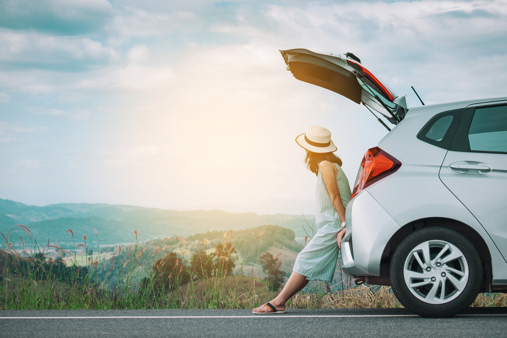 A white car is on the right side of the image, only the back half in is frame, its trunk is open and sitting in it is a woman, she is staring off into the distant hills of which there are numerous the sun is big and bright and shining