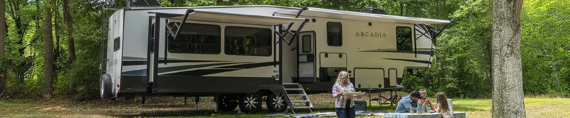 A family eating at a camping table outside a fifth wheel camper trailer at a wooded campsite.