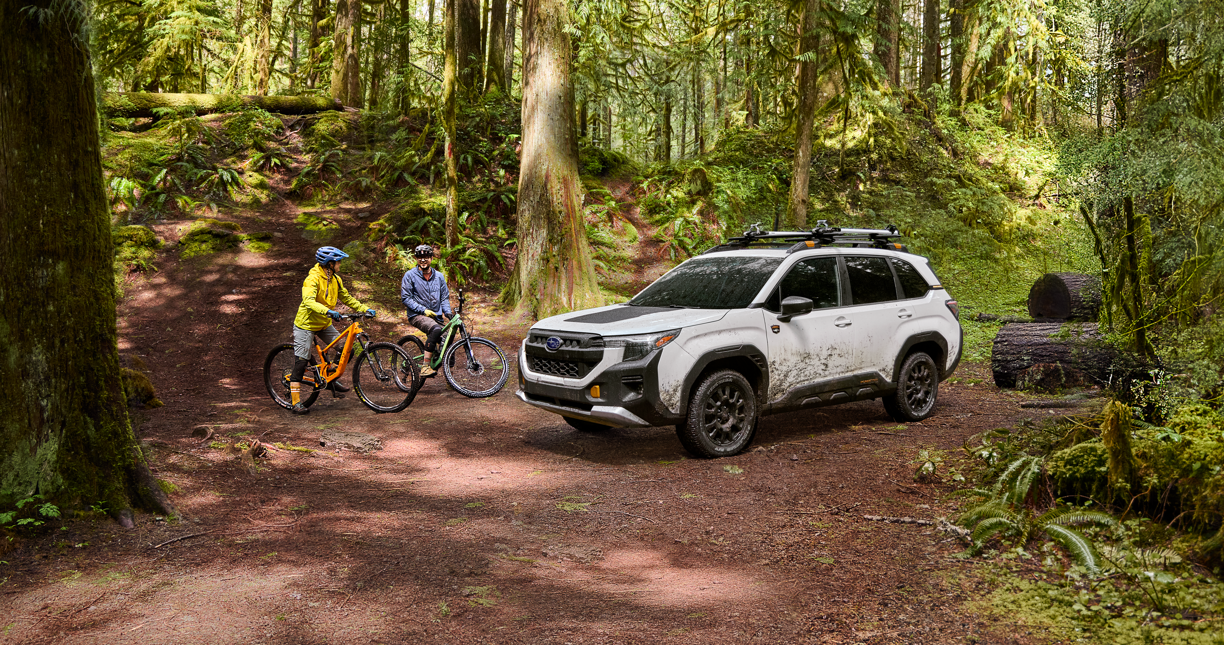 26_FORW_hero_gallery_1.png / Two people on off-road bicycles next to a muddy 2026 Subaru Forester Wilderness in white in the middle of a forest.