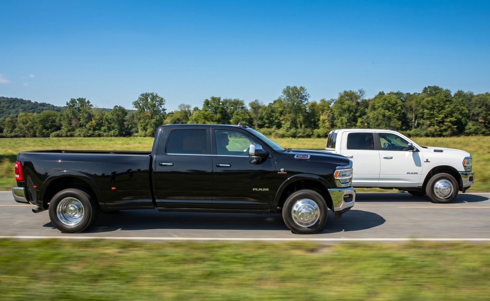 A black Ram dually pickup truck and a white Ram dually pickup truck driving side-by-side on a country road past a green field and forest on a clear day.