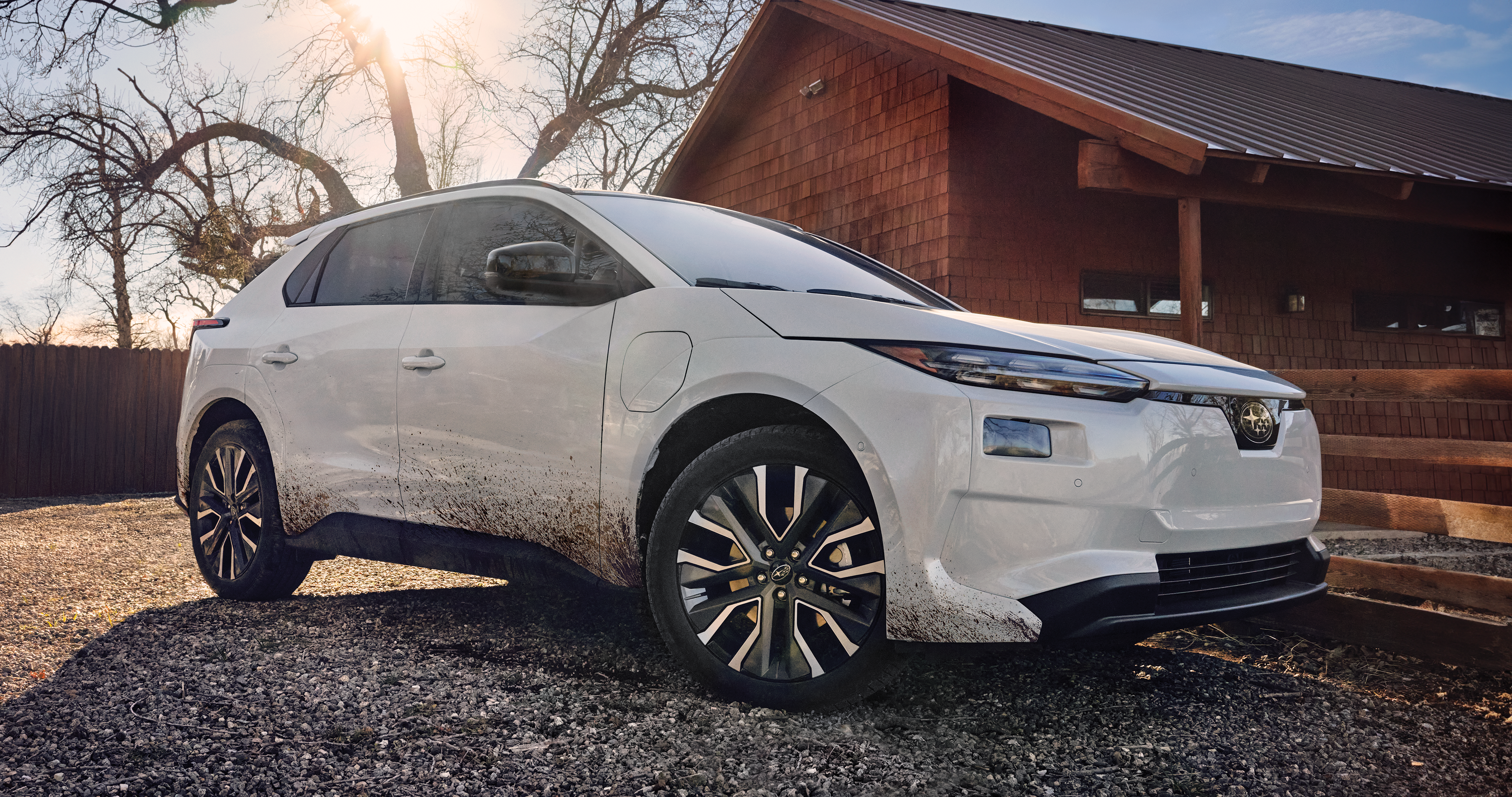 A static image of a white 2026 Subaru Solterra parked on a gravel surface beside a wooden building. 