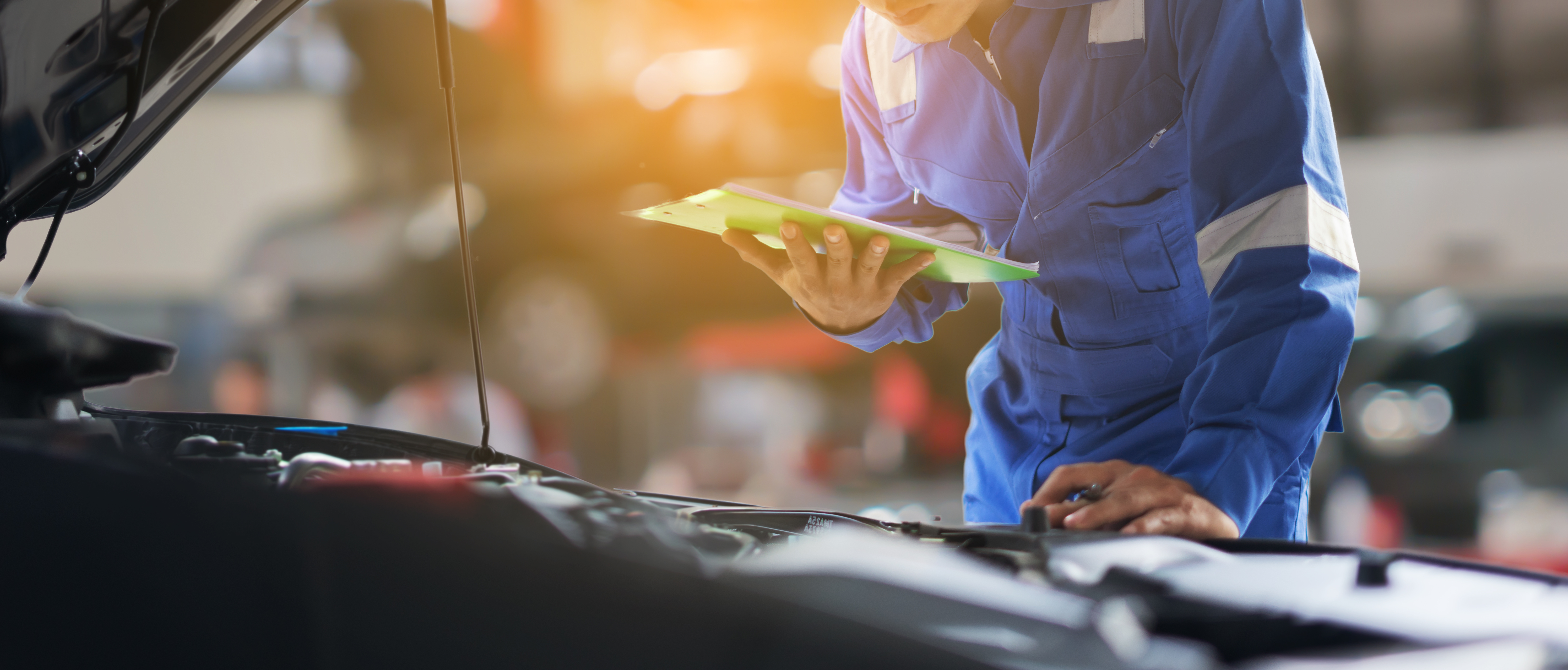 service tech with clipboard leans over open hood of vehicle in the shop