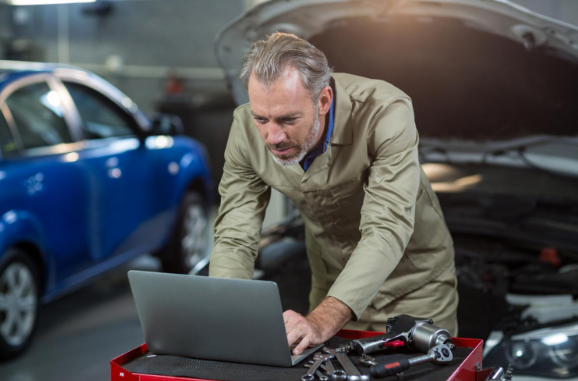 A mechanic in work clothes is bent over a laptop, likely ordering auto parts.