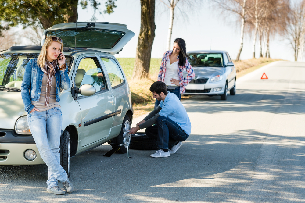 young adults parked on the side o a road fixing a flat tire