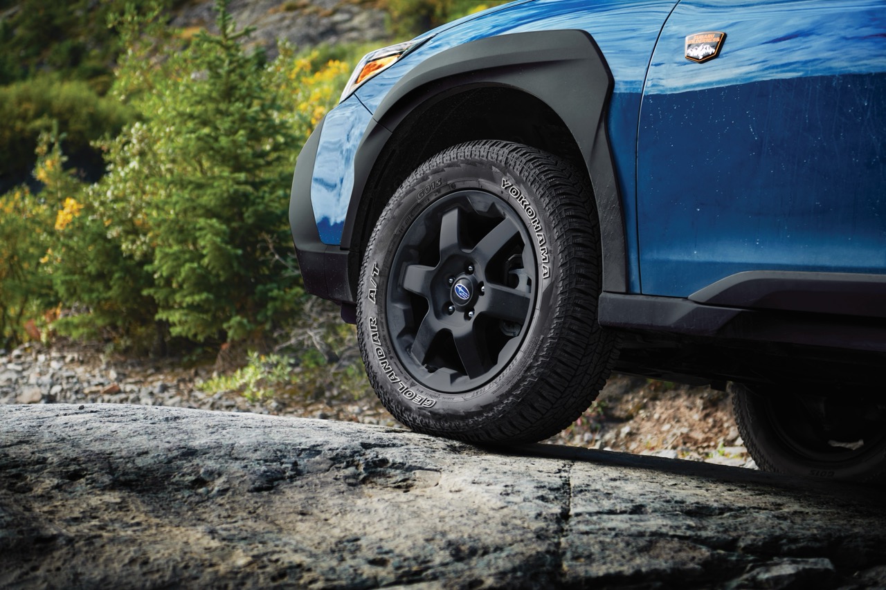A close-up static image of the front wheel and lower side panel of a blue Subaru Wilderness model parked on uneven rocky terrain. 
