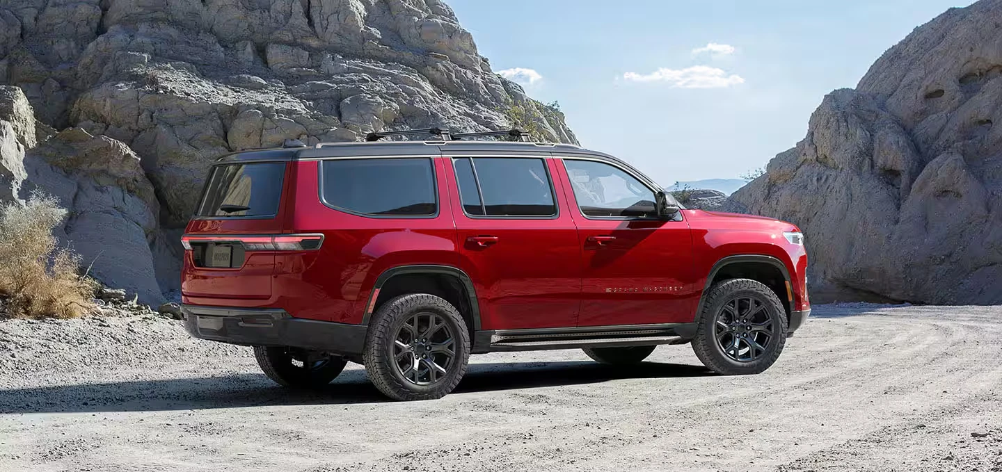 Red 2027 Jeep Grand Wagoneer SUV with black wheels, all-terrain tires and a black roof rack, seen from the rear three-quarter on a gravel desert road between pale rock outcrops under a blue sky