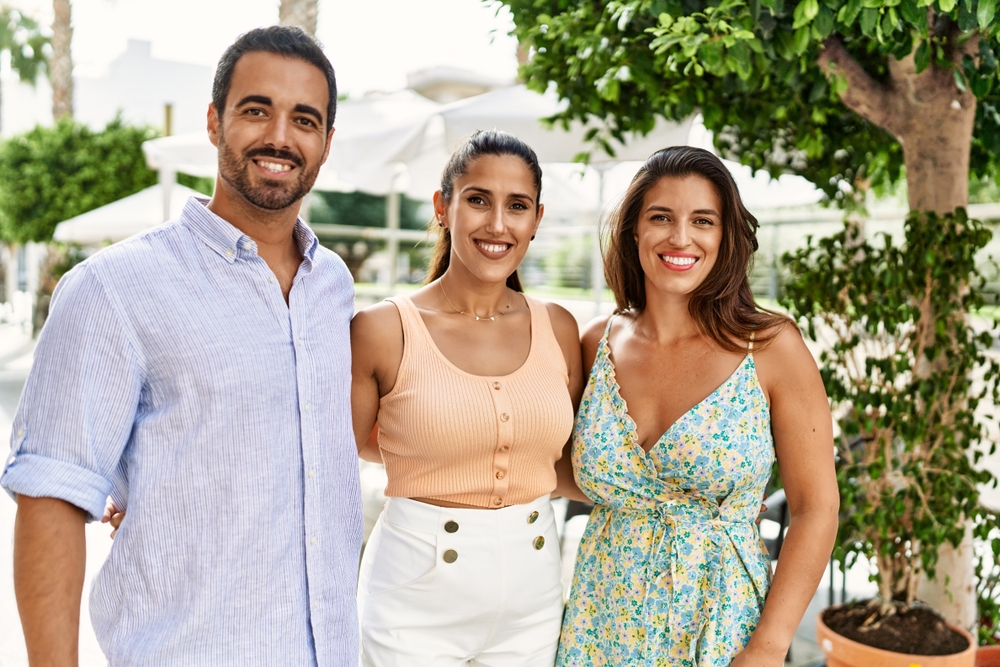 A man and two women stand side by side smiling warmly at the camera in an outdoor setting. The man wears a light blue shirt, one woman a peach tank top, and the other a floral sundress, with a leafy tree, patio umbrellas, and bright daylight behind them.