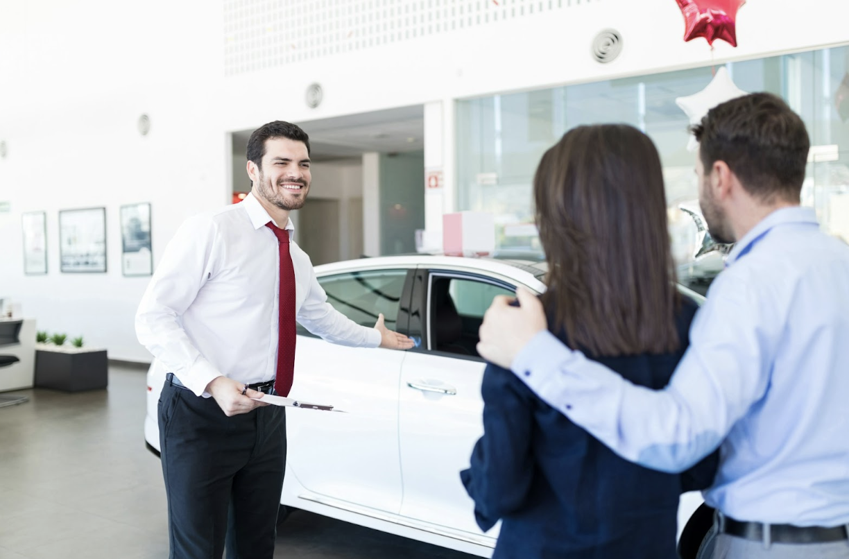 A smiling car salesman gestures toward a white car while speaking with a couple.