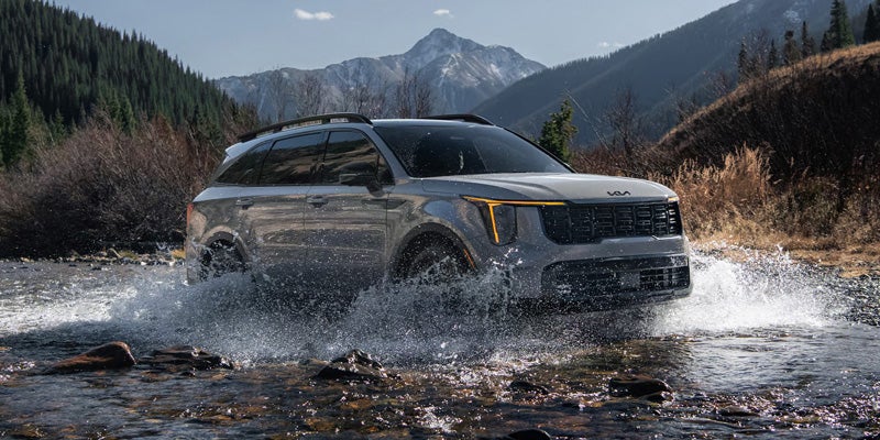 A silver 2026 Kia Sorento splashes through a rocky river with mountains in the background.