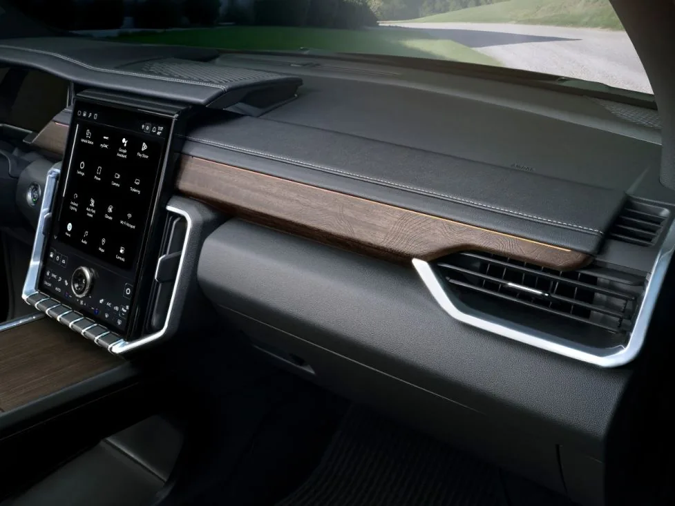 Close-up interior dashboard view of a GMC SUV showing the wood-trimmed dash panel and portrait touchscreen, with a gravel road visible through the windshield