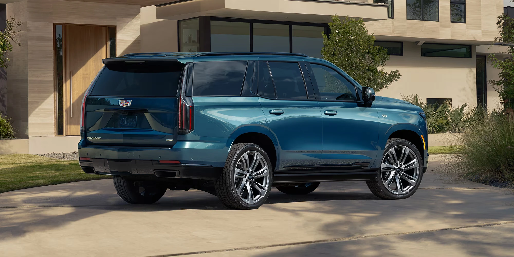 Blue Cadillac Escalade SUV seen from the rear passenger side, parked in a driveway in front of a modern house