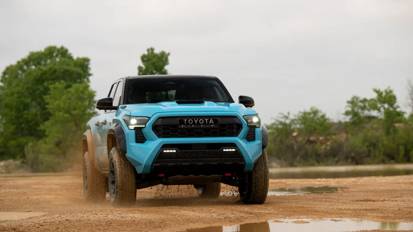 Bright blue 2026 Toyota Tacoma pickup with a TOYOTA-lettered grille, hood scoop, lower light bar and muddy off-road tires driving toward the camera across a wet dirt flat with puddles and trees behind it