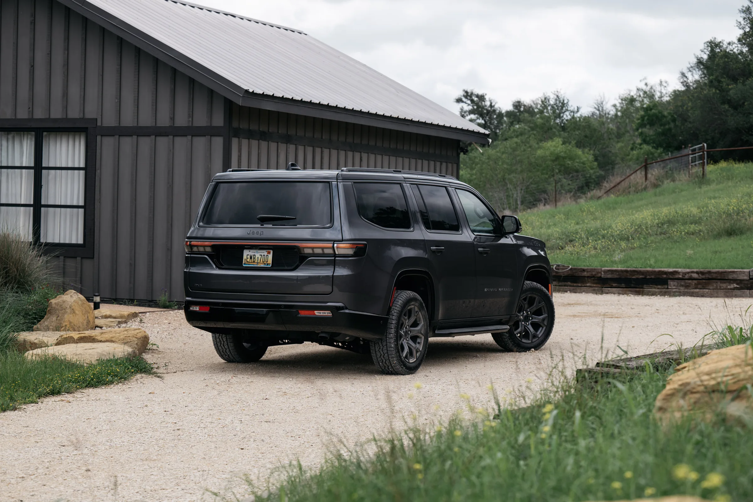 Dark gray 2026 Jeep Grand Wagoneer parked on a gravel driveway in front of a barn, shown from the rear three-quarter angle with hills in the background