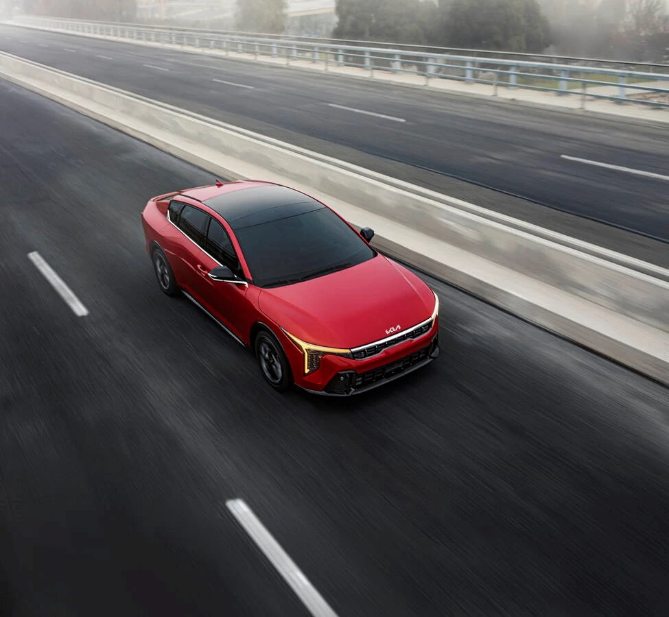 Red 2025 Kia K4 sedan seen from above driving on a foggy elevated highway, with motion blur along the pavement and guardrails.