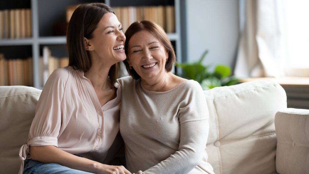 middle aged woman and her mother sitting on a couch in their living room as they laugh together