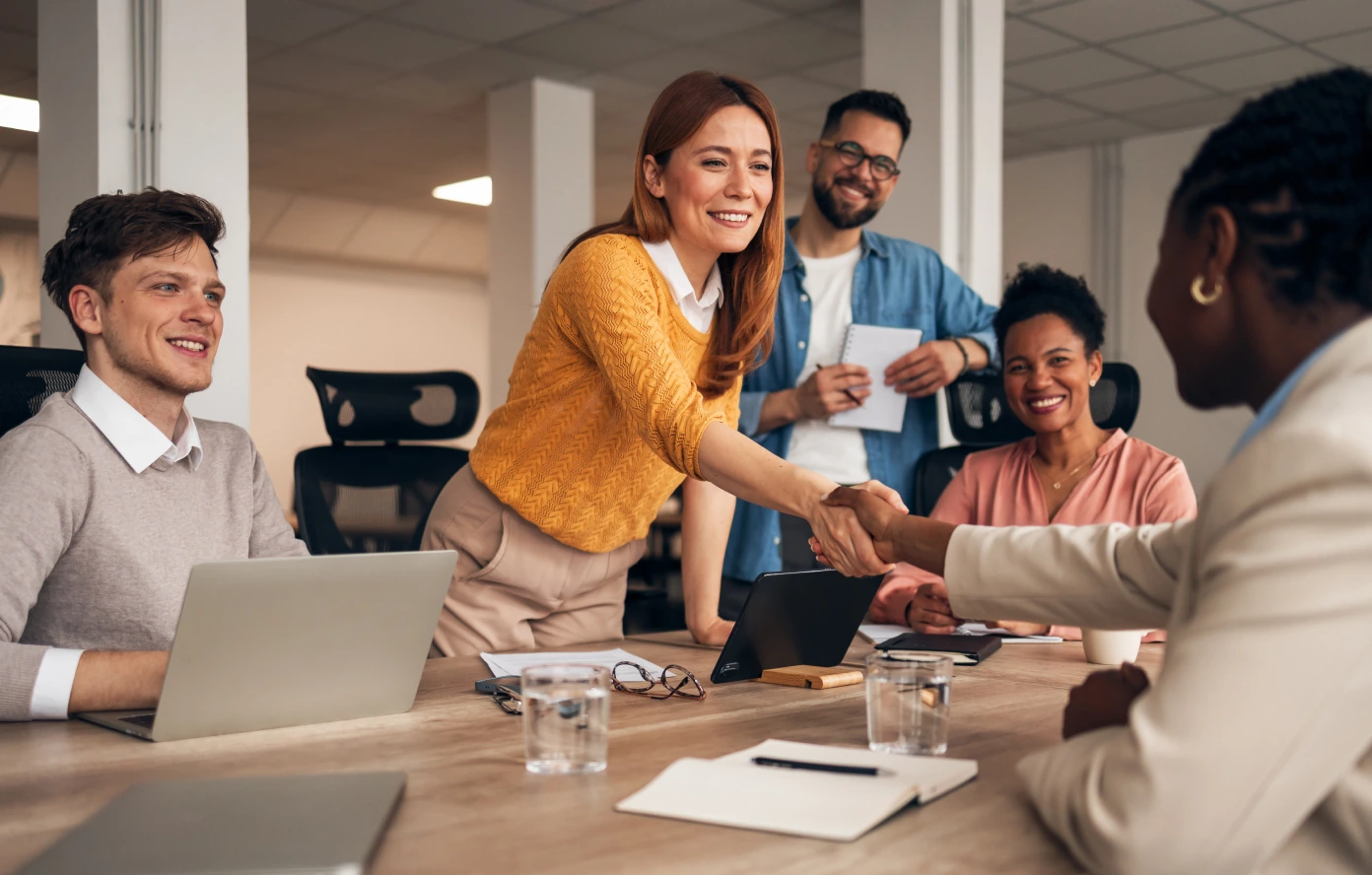 A group of people sitting around a conference table. Two women shake hands across the table.