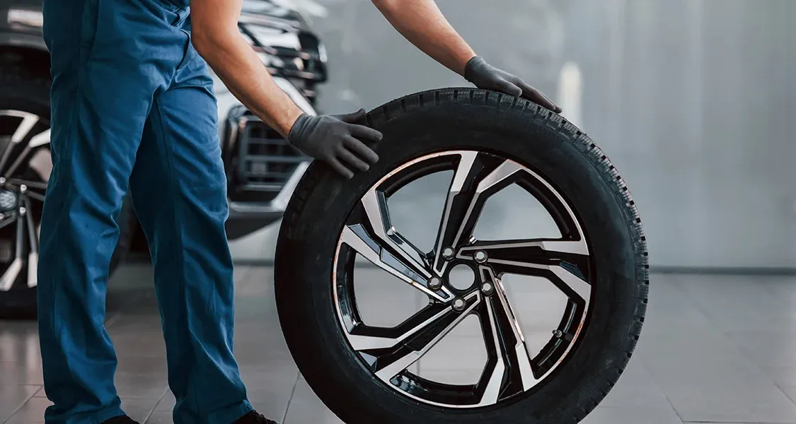 Technician wearing blue coveralls and gloves holds a tire and wheel assembly next to a dark-colored vehicle in a service bay