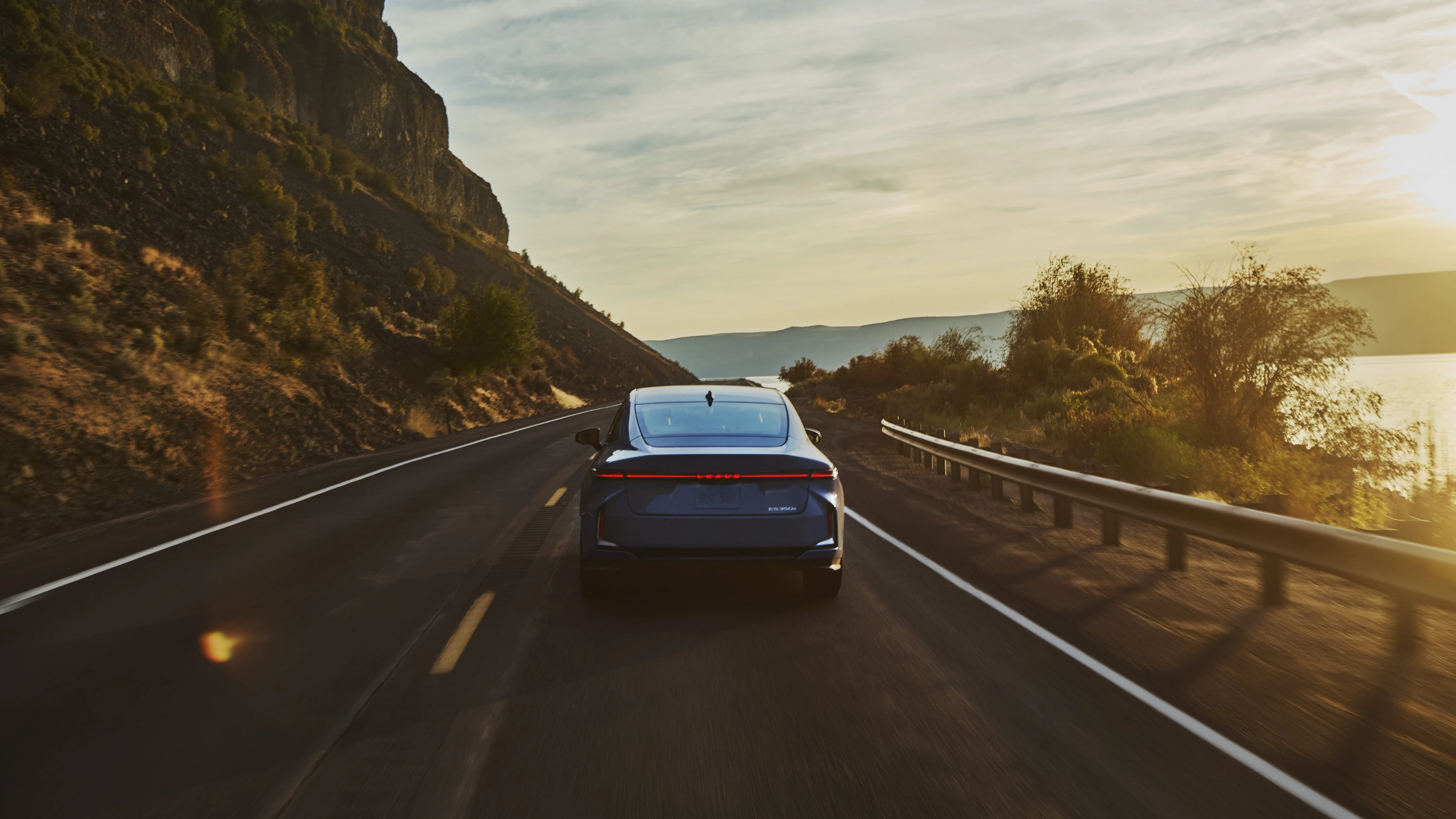 A silver Lexus sedan driving along a coastal mountain road at sunset, with cliffs rising on one side and the ocean visible on the other.