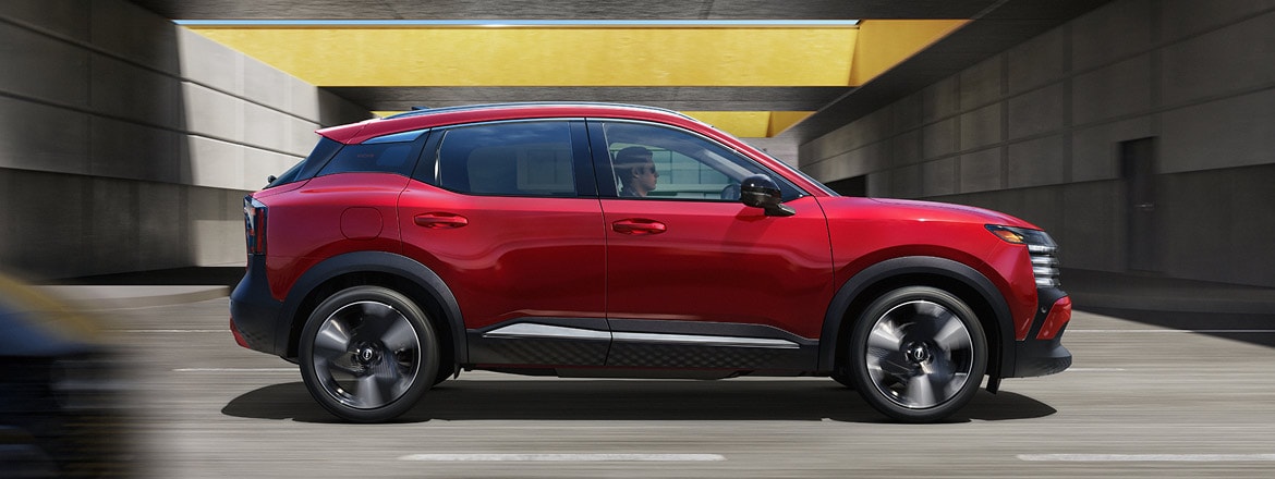 Red 2027 Nissan Kicks SUV driving through a concrete underpass with motion-blurred surroundings, shown in side profile.