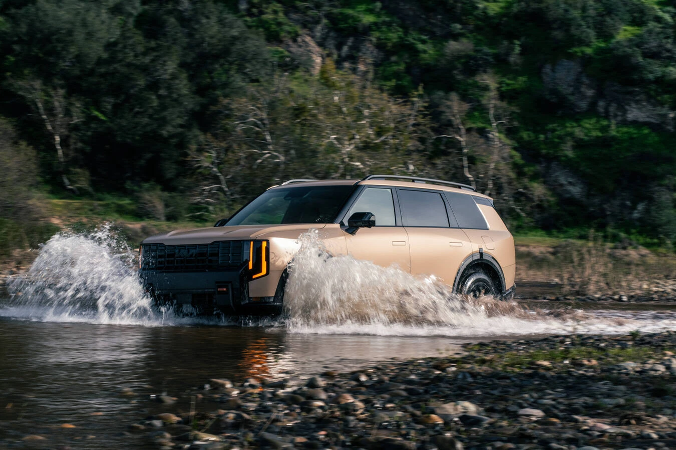 Matte tan 2027 Kia Telluride X-Pro fording a rocky stream, front three-quarter view, water splashing around the wheels with illuminated amber-accented headlights.