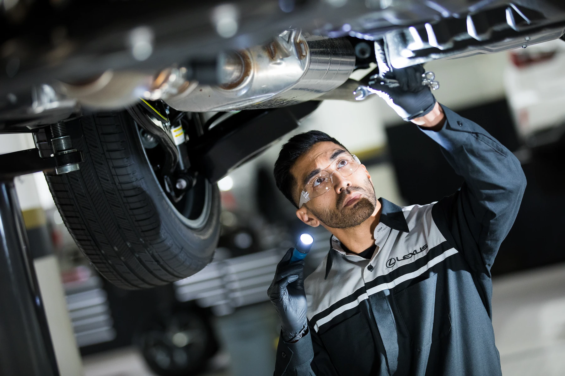 A Lexus-uniformed technician inspecting a vehicle's undercarriage on a lift with a flashlight and wrench, checking the exhaust system and suspension components.