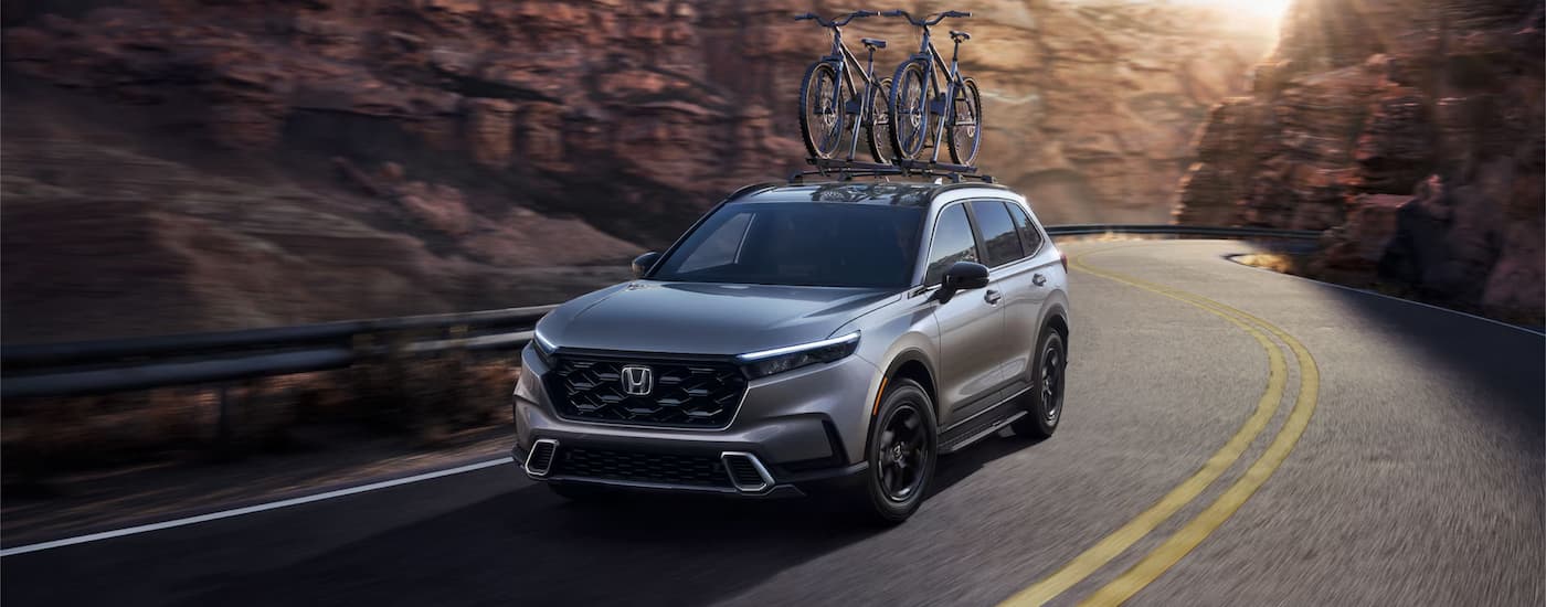 A grey 2023 Honda CR-V Sport Touring Hybrid is shown driving through a canyon with bikes on roof.