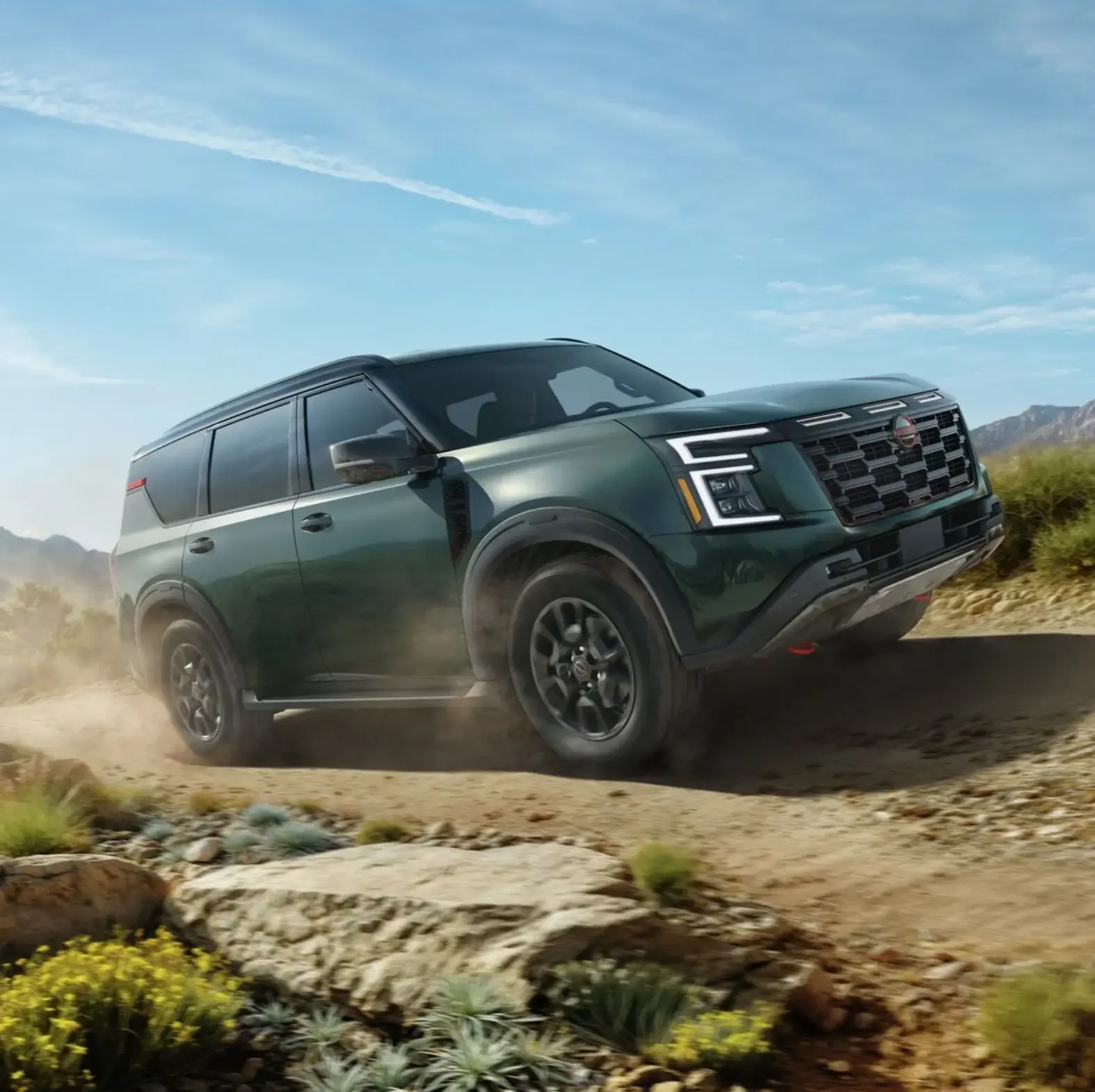 Dark green 2027 Nissan Armada with black wheels and red tow hooks kicking up dust on a desert dirt trail under a blue sky