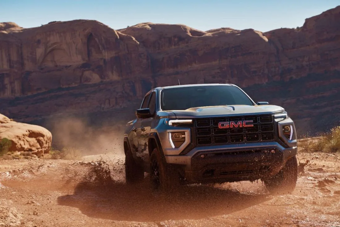 A GMC pickup truck with an off-road front bumper and light bar drives through a muddy desert canyon, kicking up dust, with red rock cliffs in the background