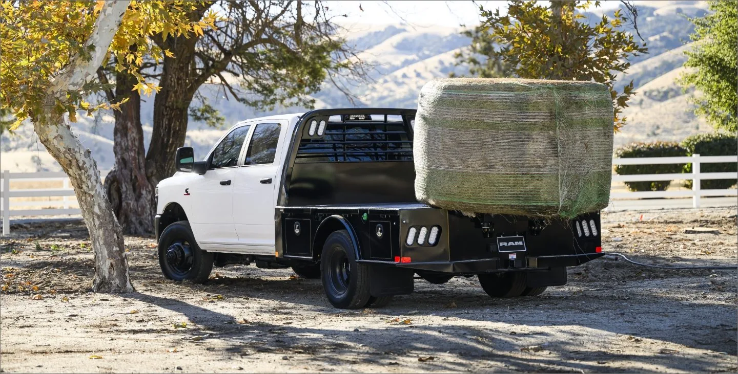 White 2026 RAM Chassis Cab with a black flatbed body hauling a large wrapped hay bale, parked under trees on a farm property with mountains in the background