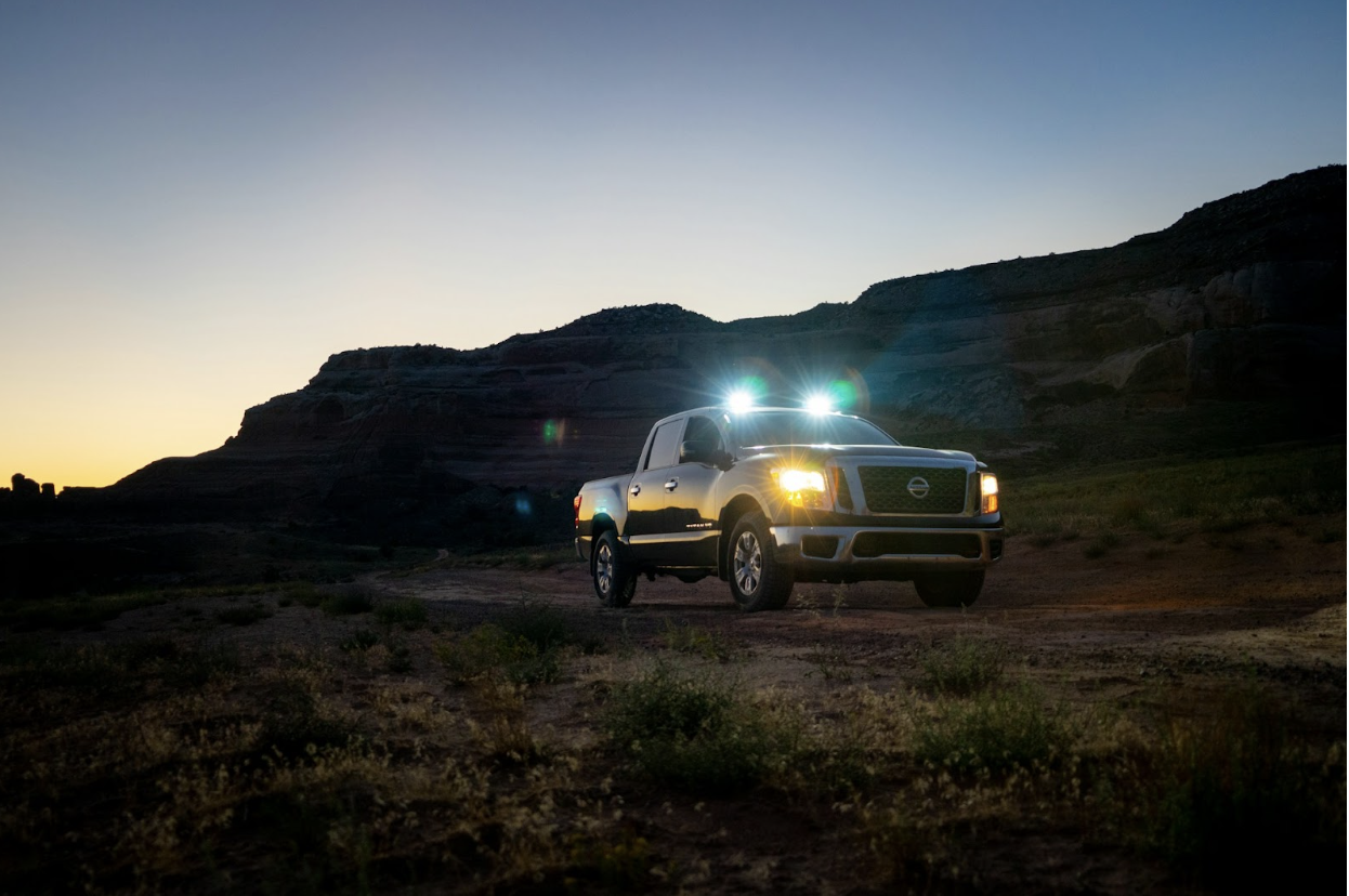 A pickup truck with bright headlights is parked on a dirt road at dusk in a desert landscape.