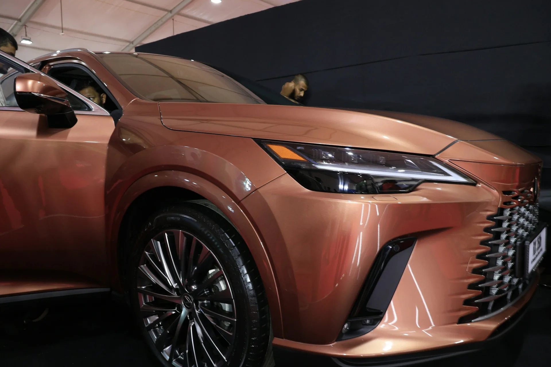  Close front three-quarter view of a copper-colored Lexus SUV on display under a white tent roof, showing its angular mesh grille, slim LED headlight and multi-spoke wheel, with two onlookers peering into and past the vehicle against a black backdrop.