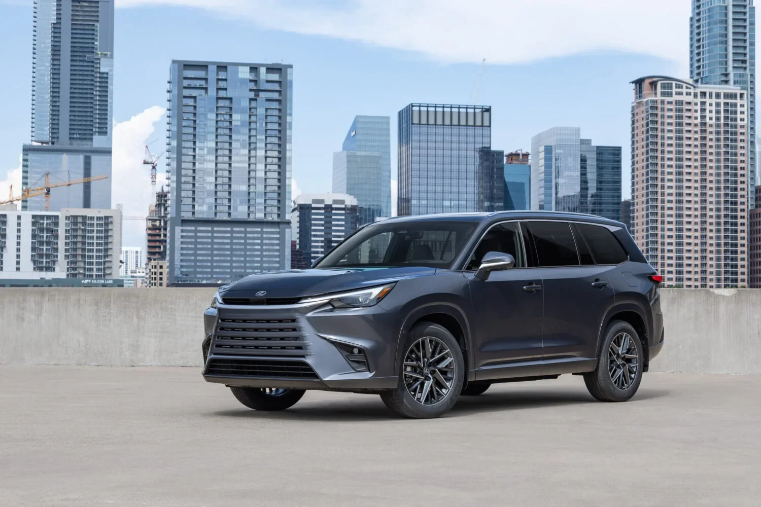 Dark gray Lexus SUV parked on a concrete rooftop deck, angled toward the left, with a skyline of glass high-rise buildings and a blue sky behind it.