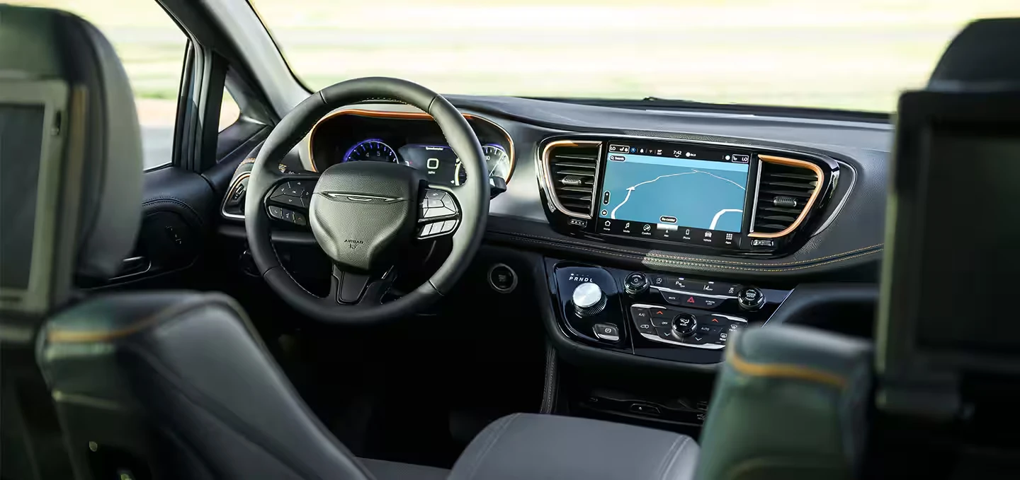 Second-row view of the 2027 Chrysler Pacifica interior with a navigation touchscreen, digital gauges, rotary shifter, orange trim and stitching, and rear-seat screens on the front headrests