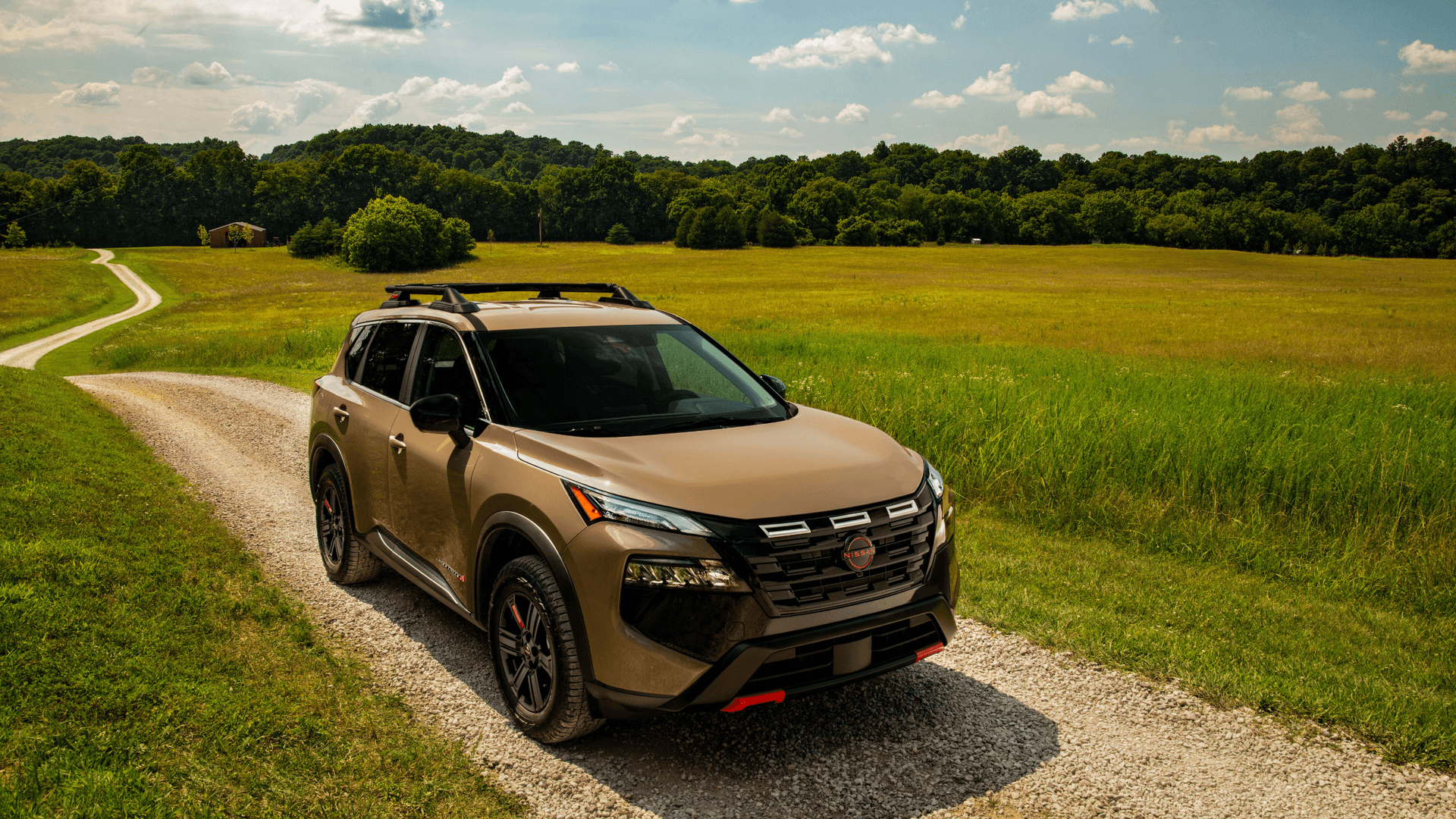 2026 Nissan Rogue® brown SUV parked on a gravel country road surrounded by open green fields and trees.