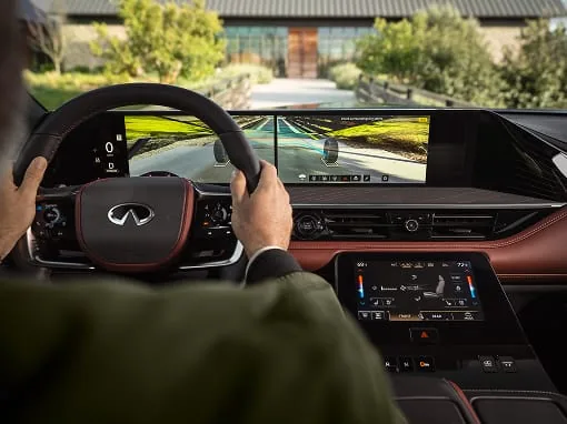 Driver’s view of the 2027 INFINITI QX80 panoramic display showing front wide-view camera assist with tire-path overlays while hands rest on the steering wheel.