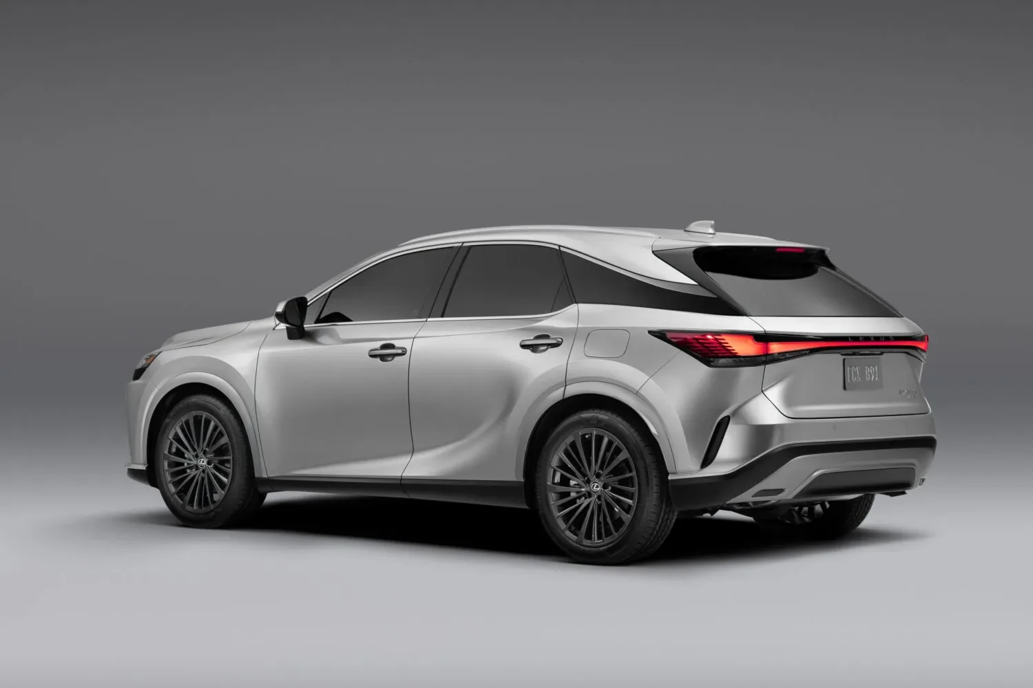 Rear three-quarter studio view of a silver Lexus SUV with a full-width red taillight bar and dark-tinted rear glass, against a gray gradient backdrop.