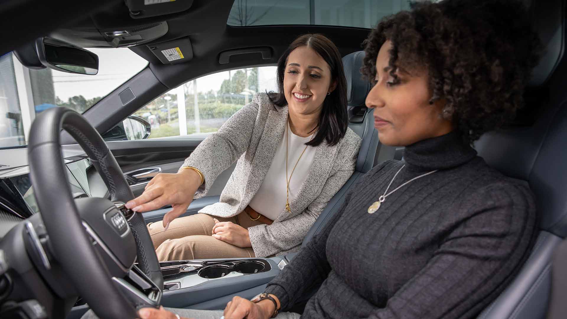 An Audi dealer explaining the features of an Audi vehicle steering wheel to a customer in the driver's seat.