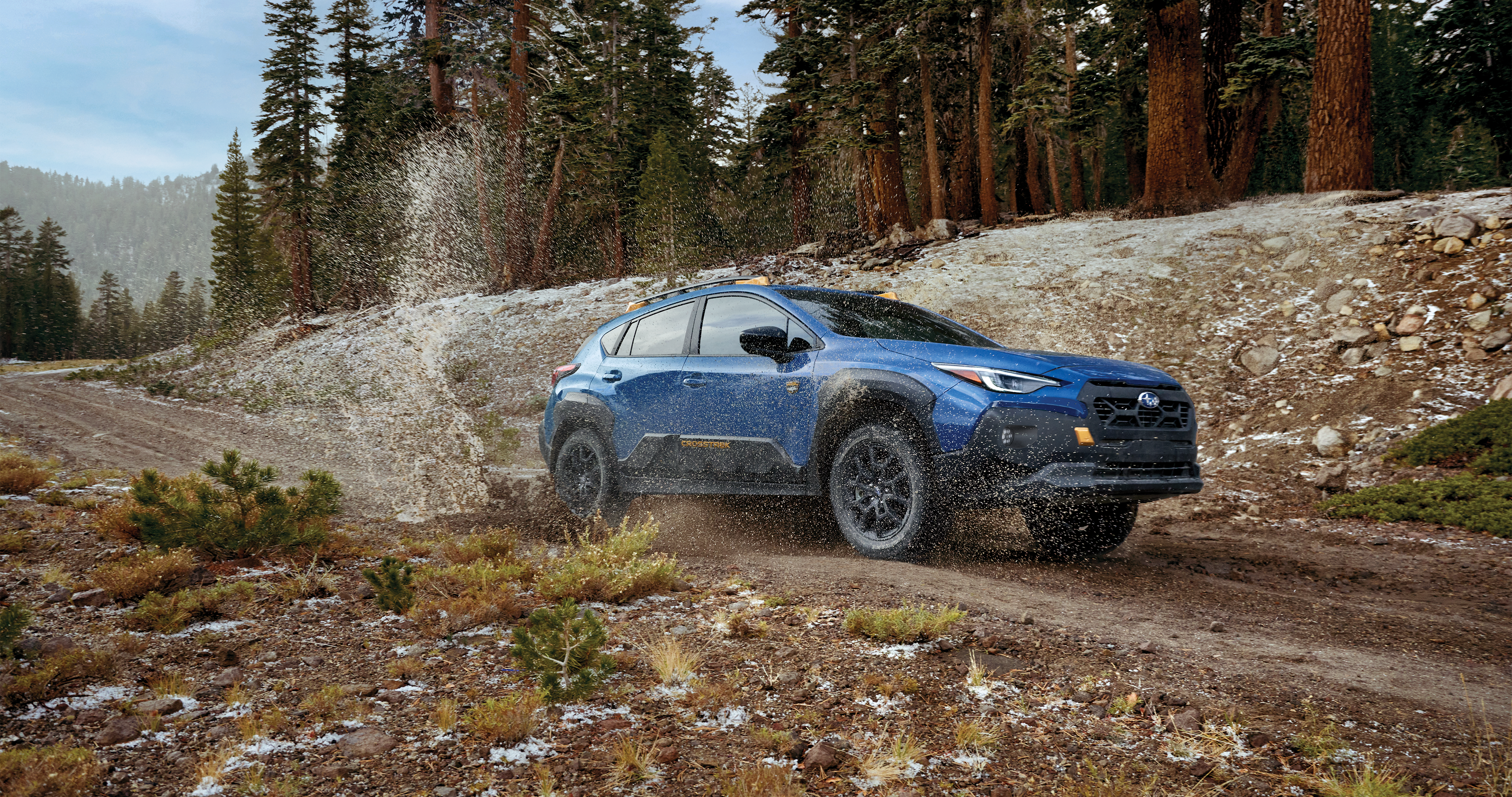 25_CTKW_hero_carousel_1.png / A Crosstrek Wilderness all-wheel drive SUV splashes through a puddle on a dirt road.