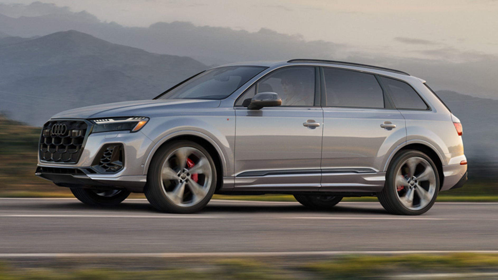 A silver Audi Q7 drives along a road with a blurred mountain backdrop.