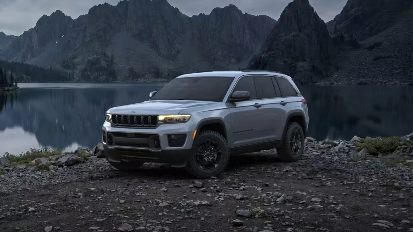 Silver 2027 Jeep Grand Cherokee SUV parked on a rocky shoreline beside a mountain lake at dusk