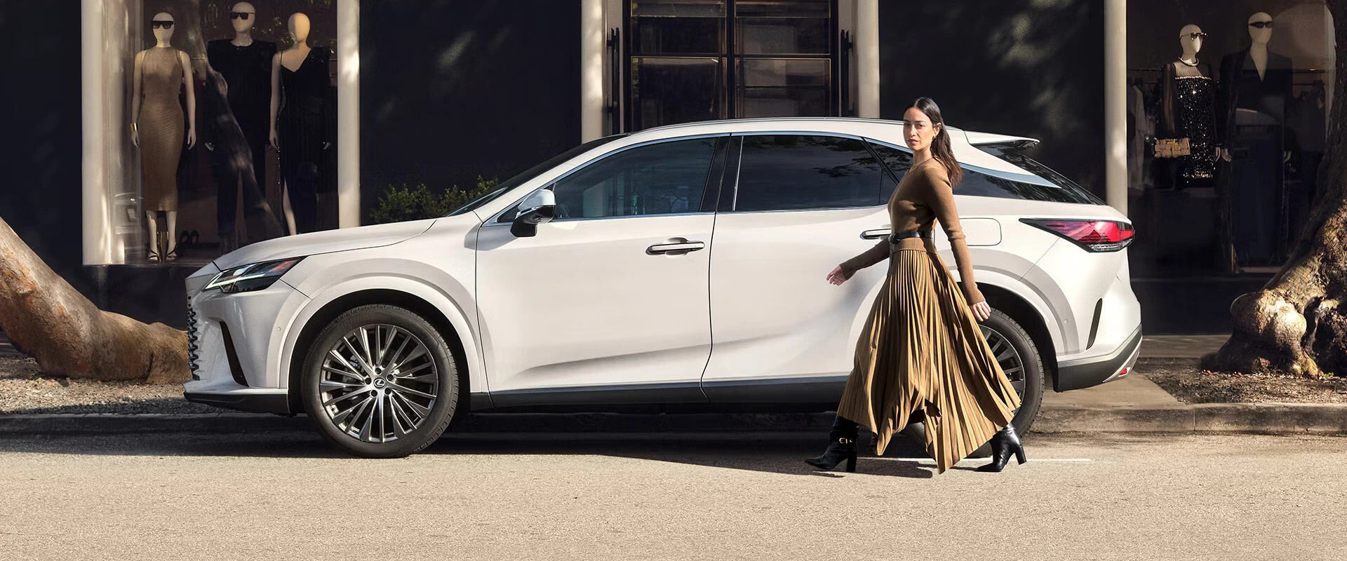 A woman in a long brown dress walks past a parked white SUV on a sunny street. Mannequins in stylish outfits are visible in a shop window behind her.