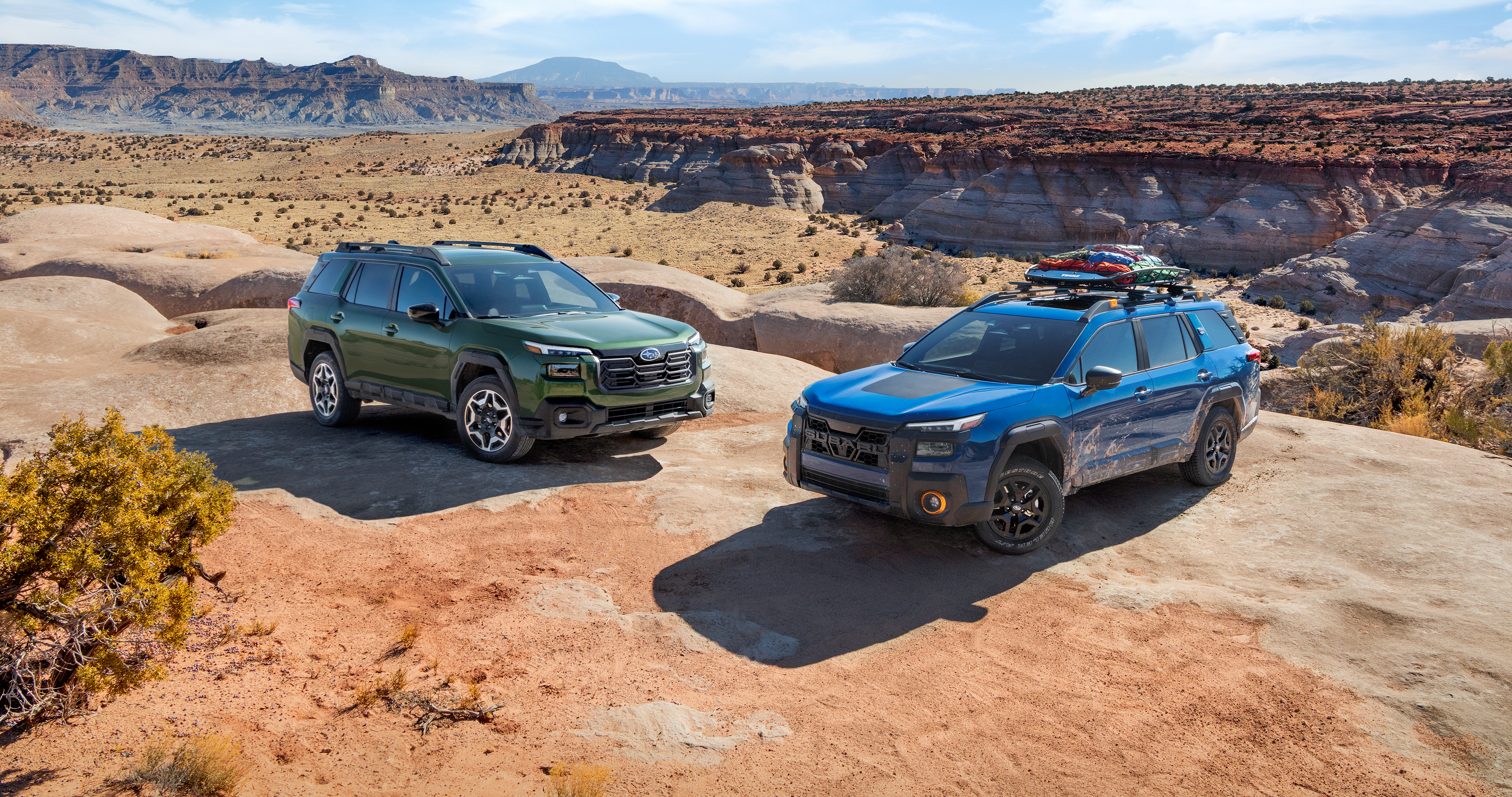 26_OBKA_hero_gallery.png / A 2026 Subaru Outback in emerald green and a 2026 Subaru Outback Wilderness in geyser blue parked next to each other on a mountain overlooking a desert. 