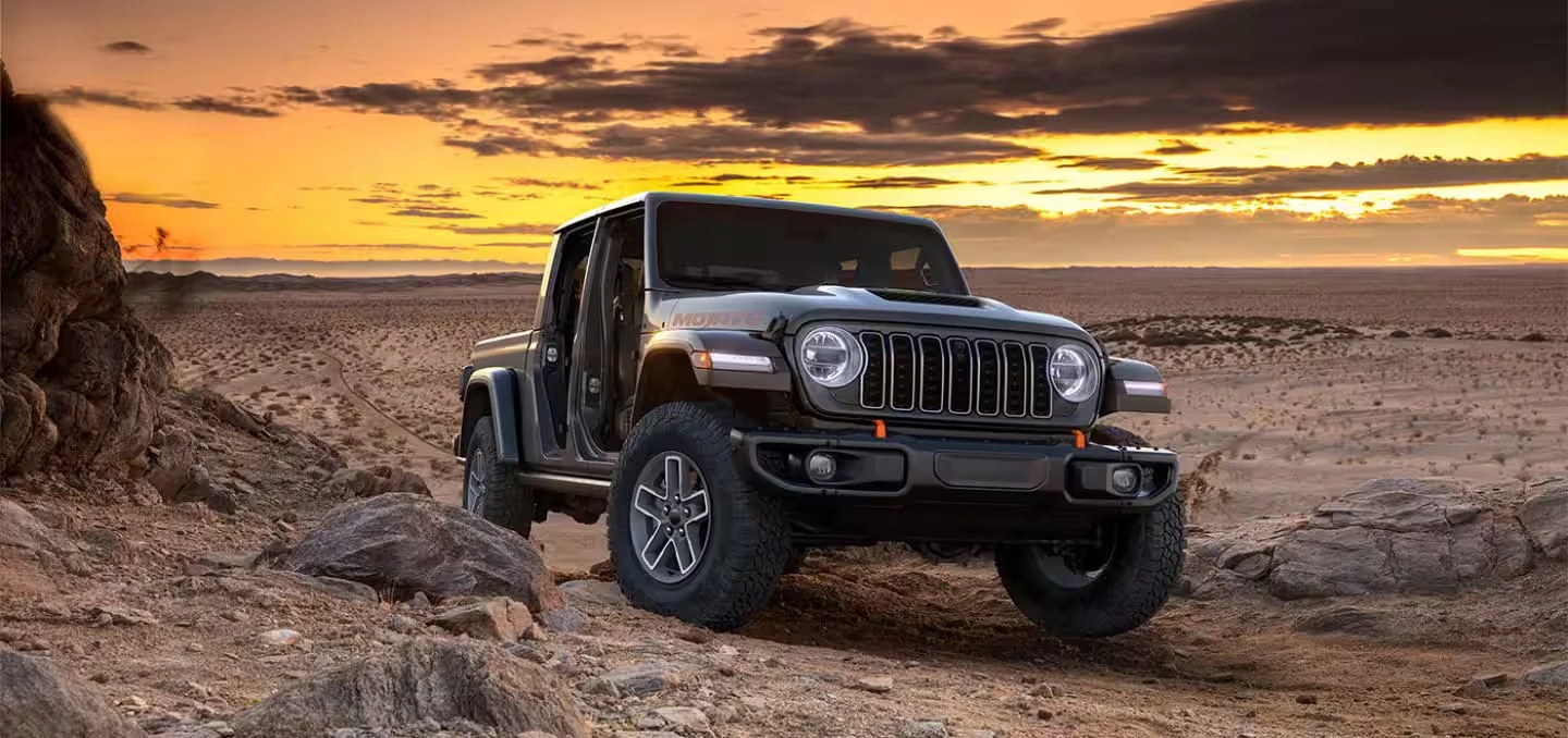Dark gray Jeep Gladiator Mojave pickup with its doors removed, black wheels and large off-road tires, parked on rocky desert terrain with a glowing orange sky and dark clouds behind it