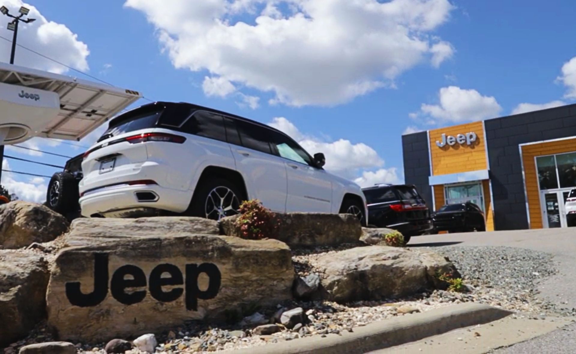 A low-angle view of a white Jeep Grand Cherokee SUV parked on an elevated rock display featuring a large "Jeep" logo engraved on the main stone, positioned outside a modern dealership building with wood-paneled accents under a blue sky with white clouds.