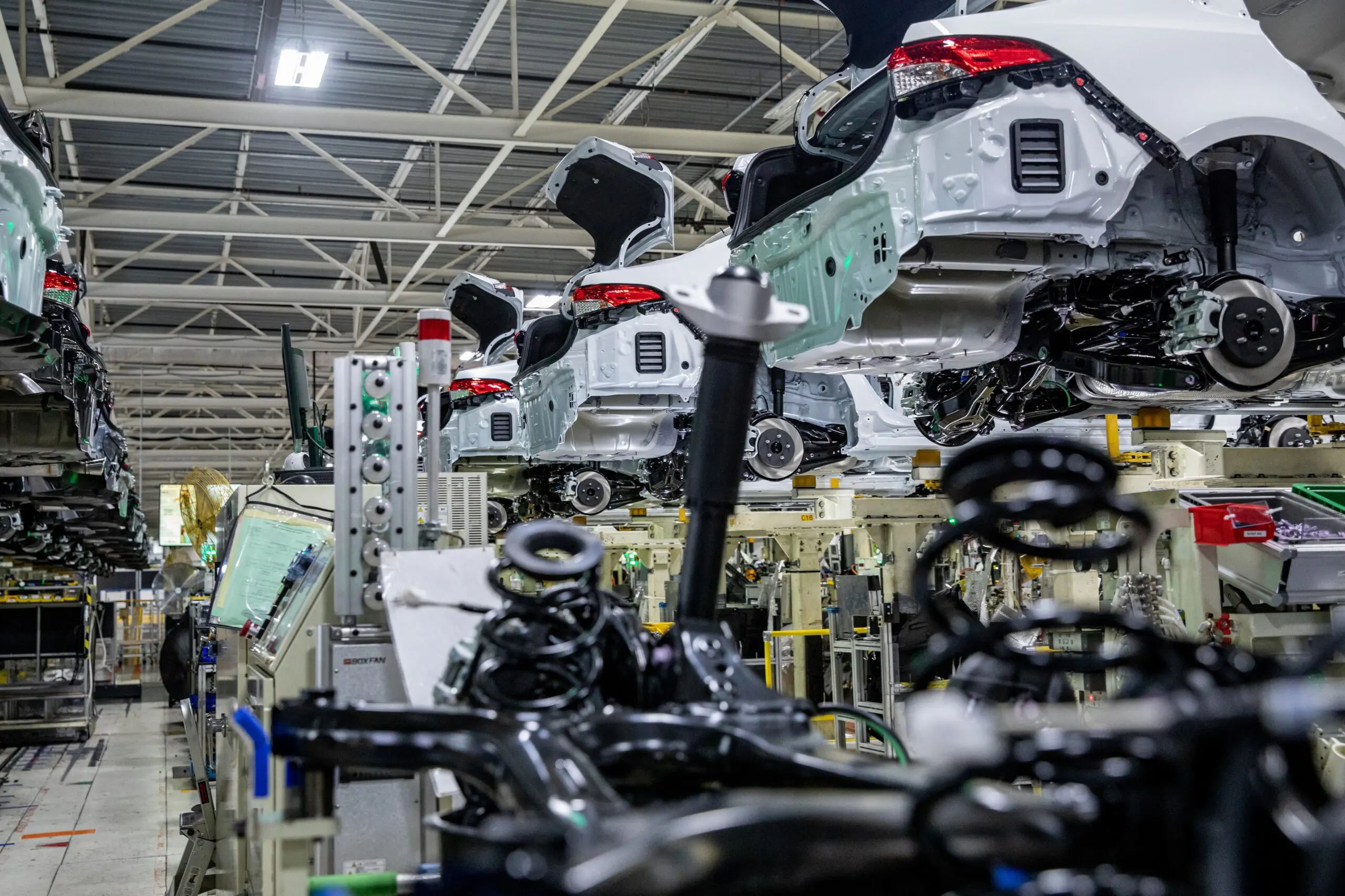 Multiple Toyota vehicles move along an assembly line inside a U.S. manufacturing plant, with exposed frames and components visible during the production process.