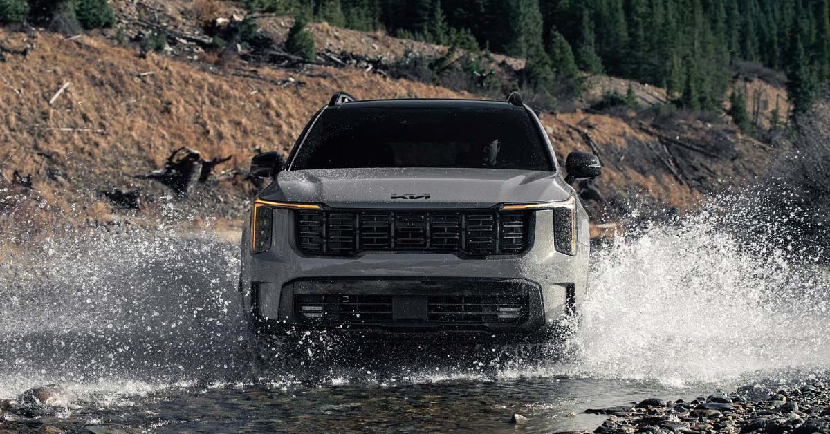 A front-facing view of a grey 2026 Kia Sorento, driving through a shallow, rocky riverbed and splashing water high on both sides