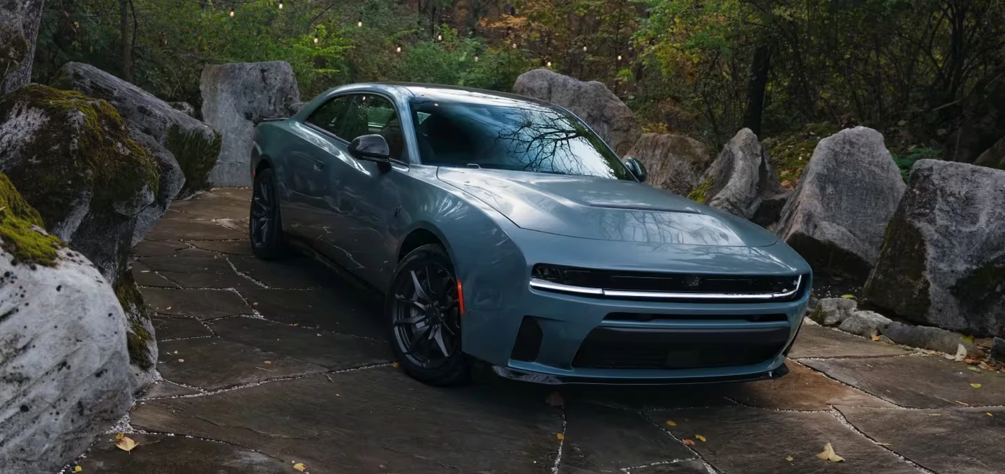 Blue-gray 2027 Dodge Charger with a full-width illuminated front light bar and black wheels, parked on a flagstone patio surrounded by mossy boulders and autumn foliage