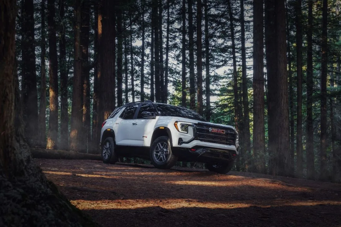 White GMC SUV with an AT4 badge driving through a sunlit pine forest, kicking up dust on a dirt trail